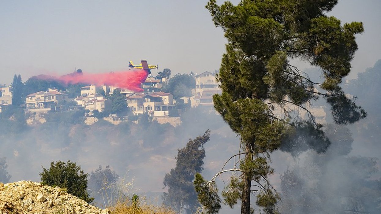 Airplane dropping fire retardant on a wildfire with homes on a hillside in the background.