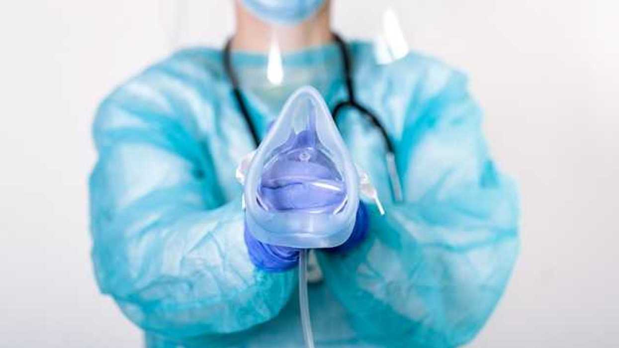 An anesthesiologist wearing a blue jumpsuit and face mask holding an anesthesia mask for a patient