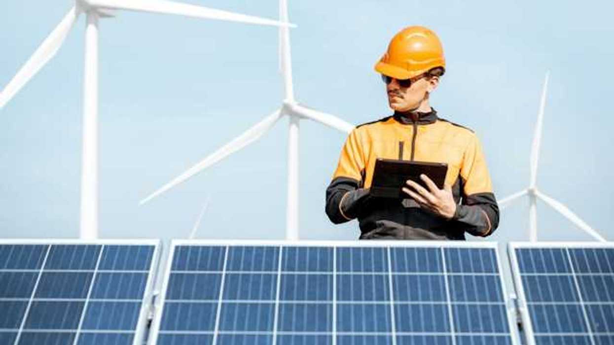 An energy worker examining solar panels with wind turbines in the background