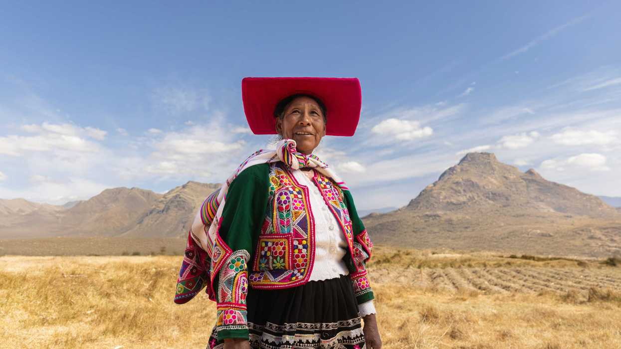 An Indigenous woman with a red hat and colorful traditional dress standing in a golden field with mountains in background..