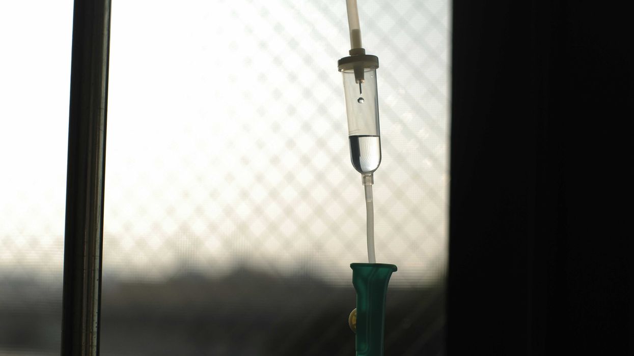 An IV drip with tube illuminated in front of a window with a blurred landscape in background.