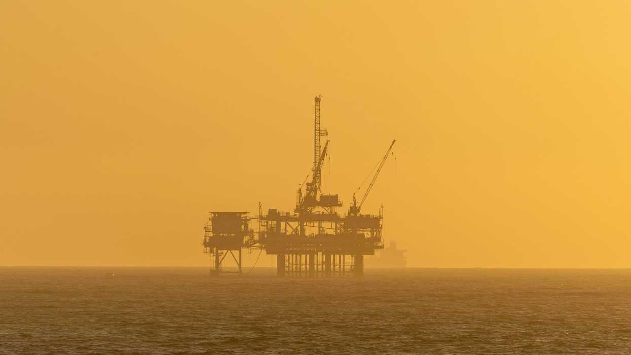 An oil rig in silhouette against a warm orange sky over the ocean.