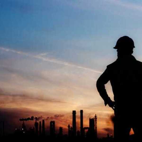 An oil worker silhouetted against a darkening sky and polluting smokestacks in the background