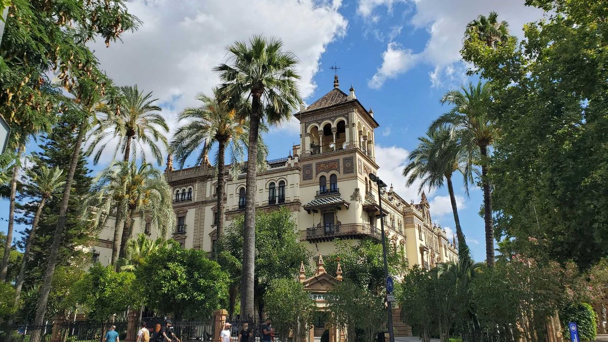 An old Spanish building surrounded by palms and other green trees with people walking in front.