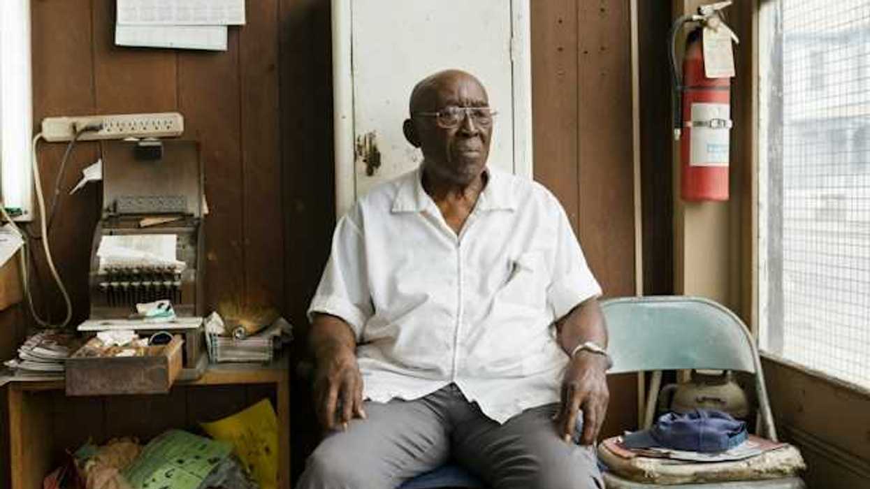 An older Black man sitting on a chair in a cluttered room
