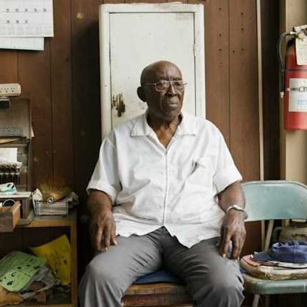 An older Black man sitting on a chair in a cluttered room