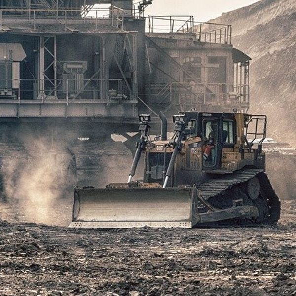 An open pit mine with dust kicked up by equipment and a processing building in the background.