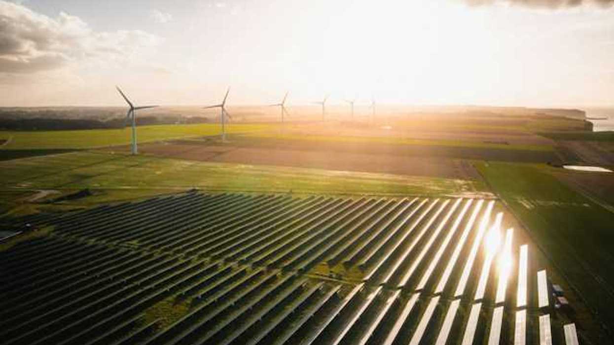 An overhead view of rows of solar panels and wind turbines in the distance