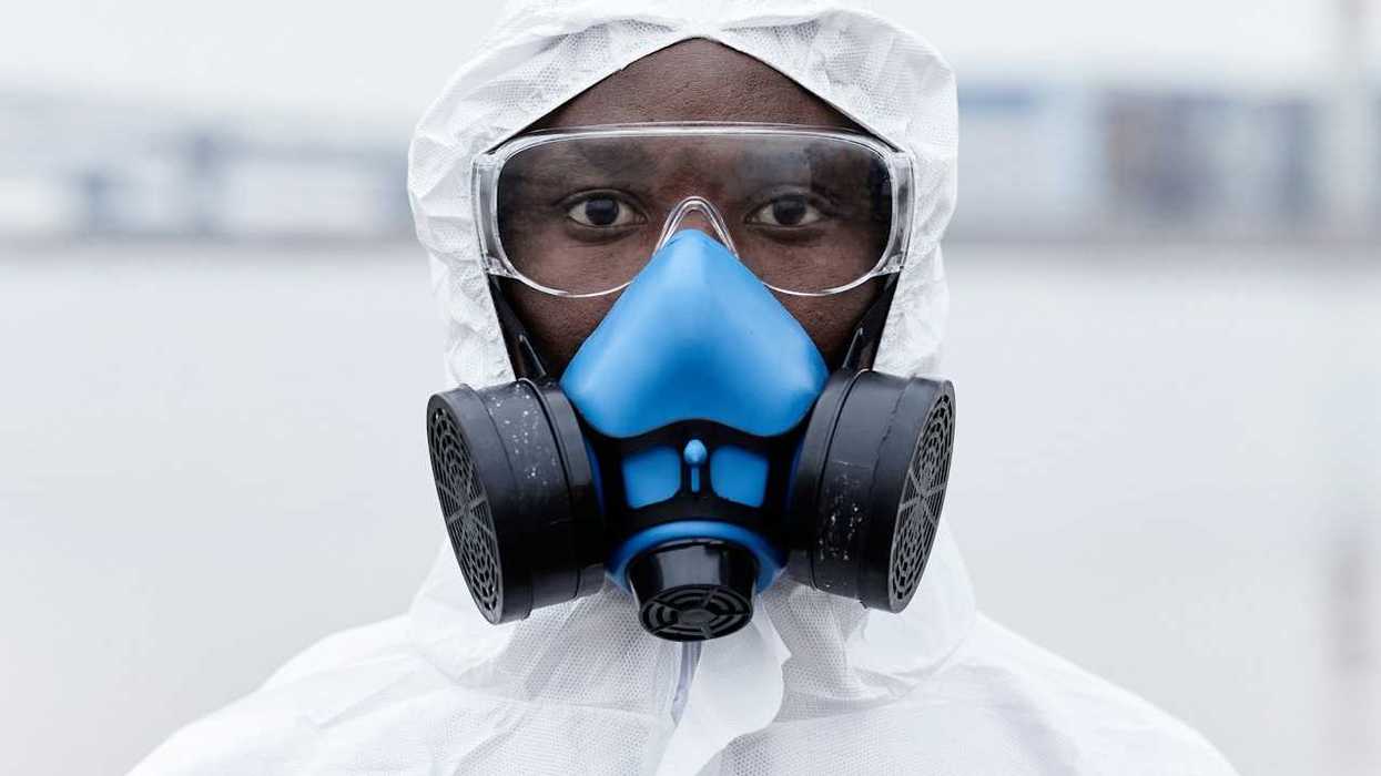 Black health care worker in protective gear—blue/black respirator, white protective suit, safety glasses.