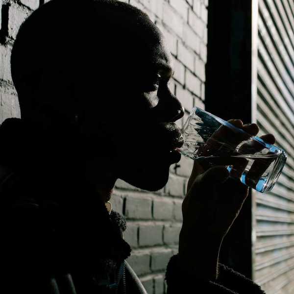 Black man leaning against brick wall drinking from a water glass