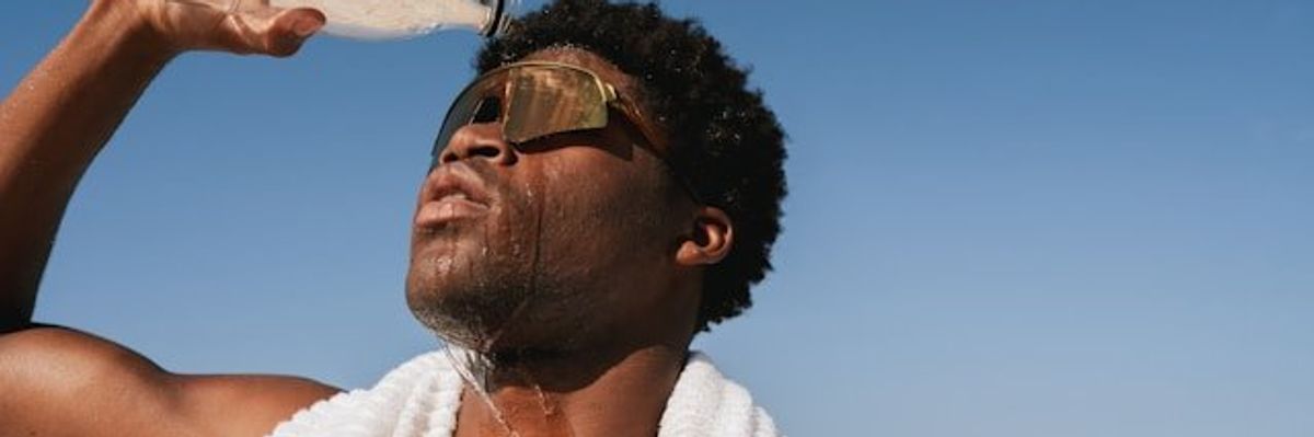 Black man pouring water over his head on a hot day with blue ocean in background.
