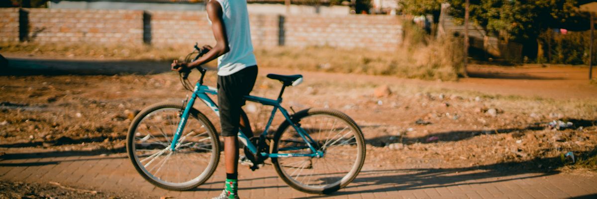 Black youth straddling blue bicycle on a sidewalk through a vacant lot with homes in background.