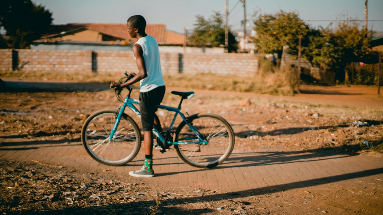 Black youth straddling blue bicycle on a sidewalk through a vacant lot with homes in background.