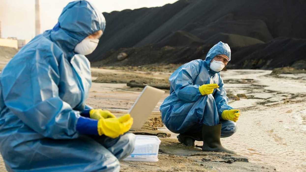 Blue-suited, masked, technicians sampling soil and water adjacent to a large pile of toxic cola ash