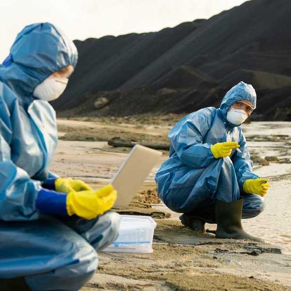 Blue-suited, masked, technicians sampling soil and water adjacent to a large pile of toxic cola ash