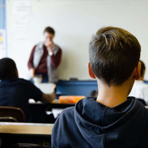 boy in black hoodie sitting on chair in classroom with other students, facing a teacher.