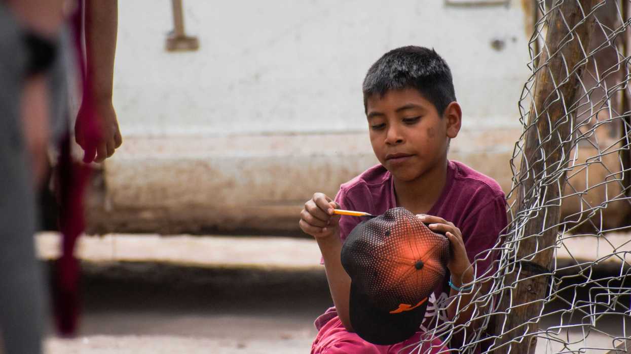 boy in red t-shirt holding baseball cap by a chain link fence.