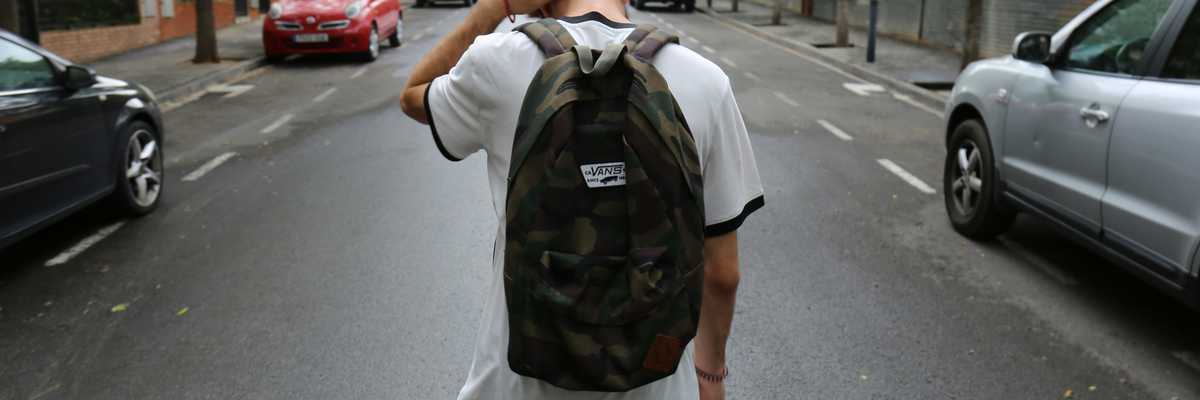 boy wearing white shirt and black shorts carrying backpack standing on black concrete road between vehicles and trees during daytime