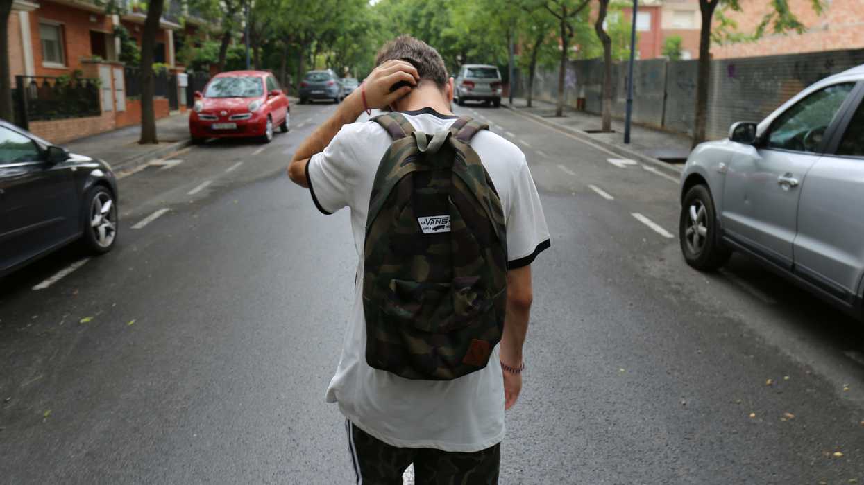 boy wearing white shirt and black shorts carrying backpack standing on black concrete road between vehicles and trees during daytime