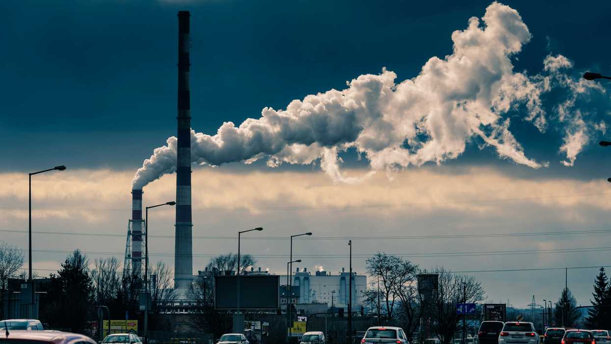 Cars driving on a street with a factory smokestack polluting in the background.