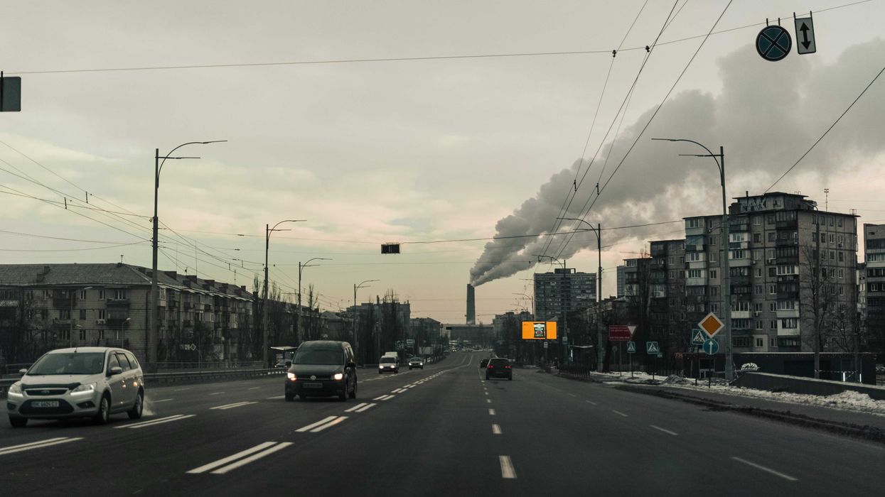 Cars on a road flanked by apartment buildings with a polluting smokestack in background.