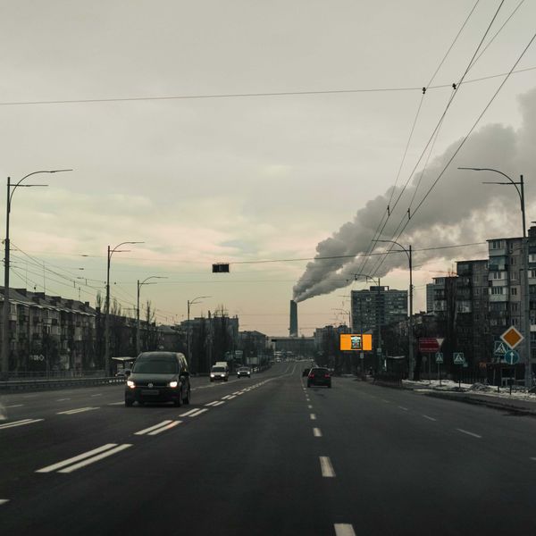 Cars on a road flanked by apartment buildings with a polluting smokestack in background.