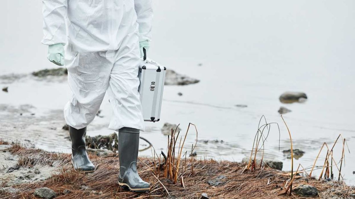 Chest-down person in white protective suit and rubber boots carrying metal case, walking a shoreline.