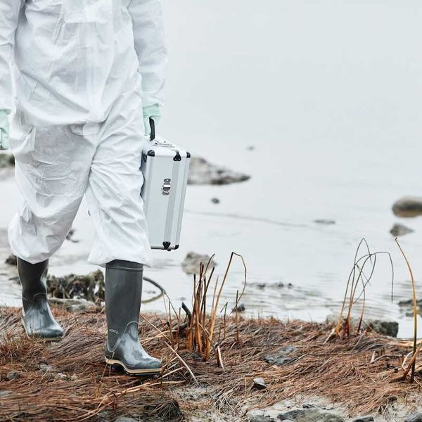 Chest-down person in white protective suit and rubber boots carrying metal case, walking a shoreline.