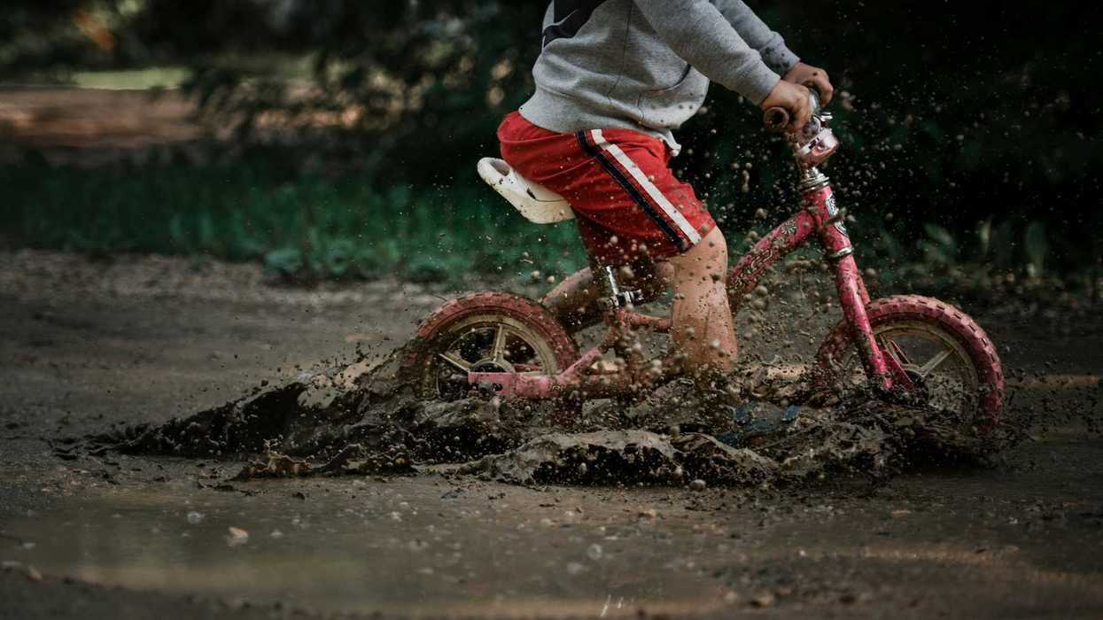 child in gray jacket and red pants riding red bicycle on flooded muddy road.