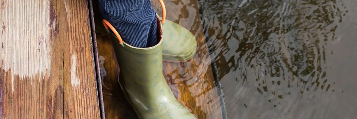 Child's legs and feet in green rubber boots stands on flooded wooden steps.