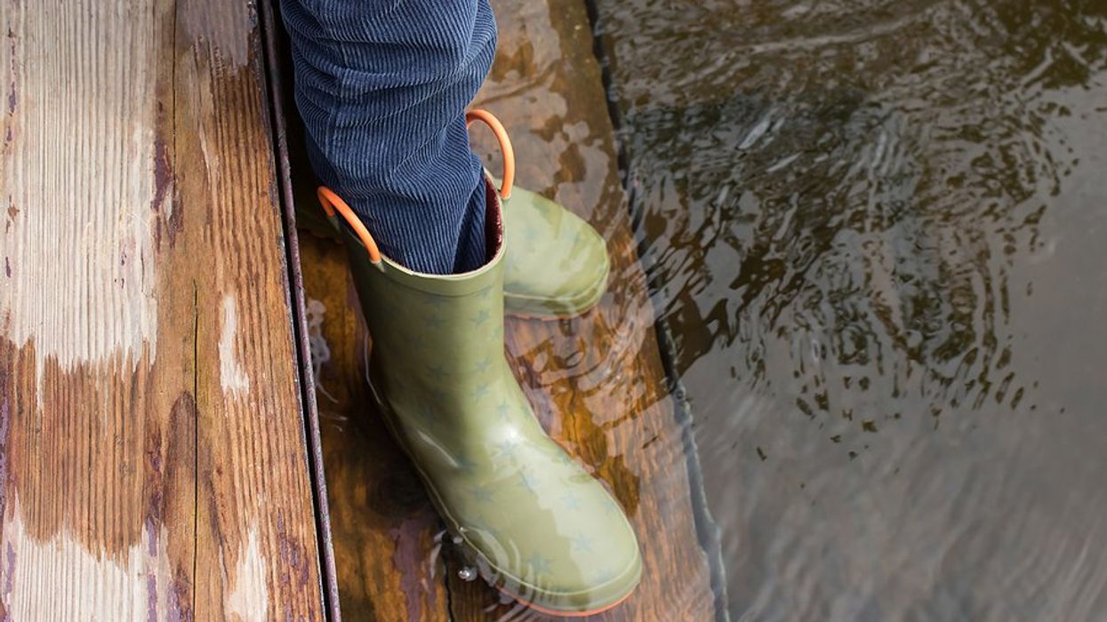 Child's legs and feet in green rubber boots stands on flooded wooden steps.