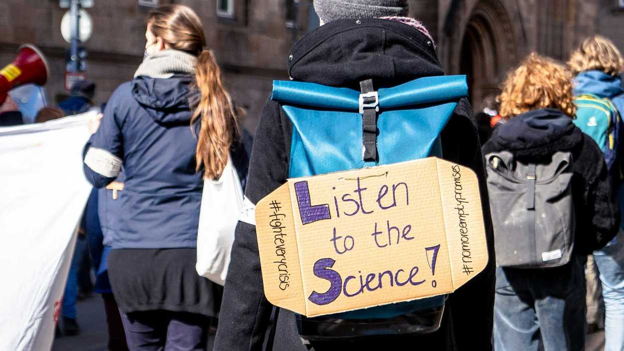 climate demonstrator in a crowd wearing cardboard sign on back that reads "listen to the science!")