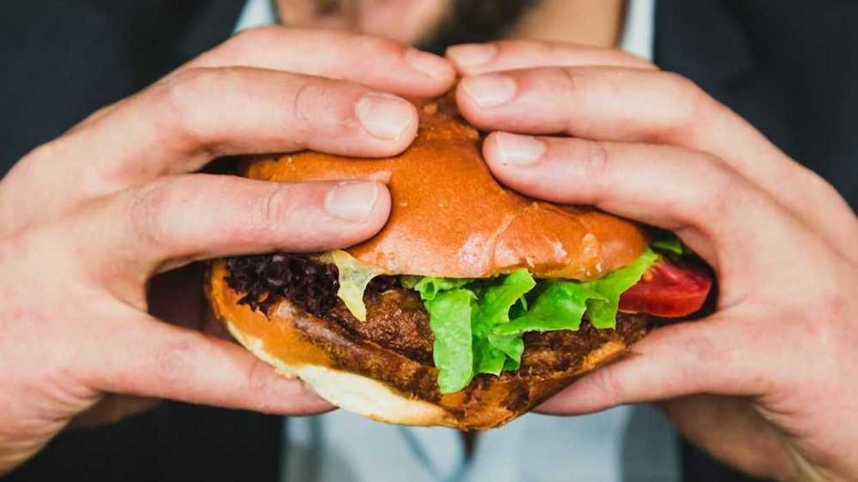 Closeup of man with a two-handed grip on a hamburger in a bun, mouth open, about to eat.