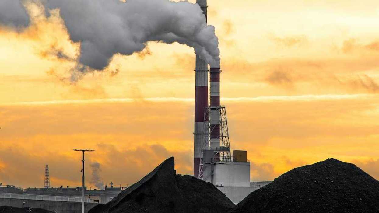 Coal fired power plant with piles of coal beneath red & white stacks emitting smoke