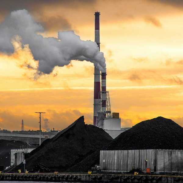 Coal fired power plant with piles of coal beneath red & white stacks emitting smoke