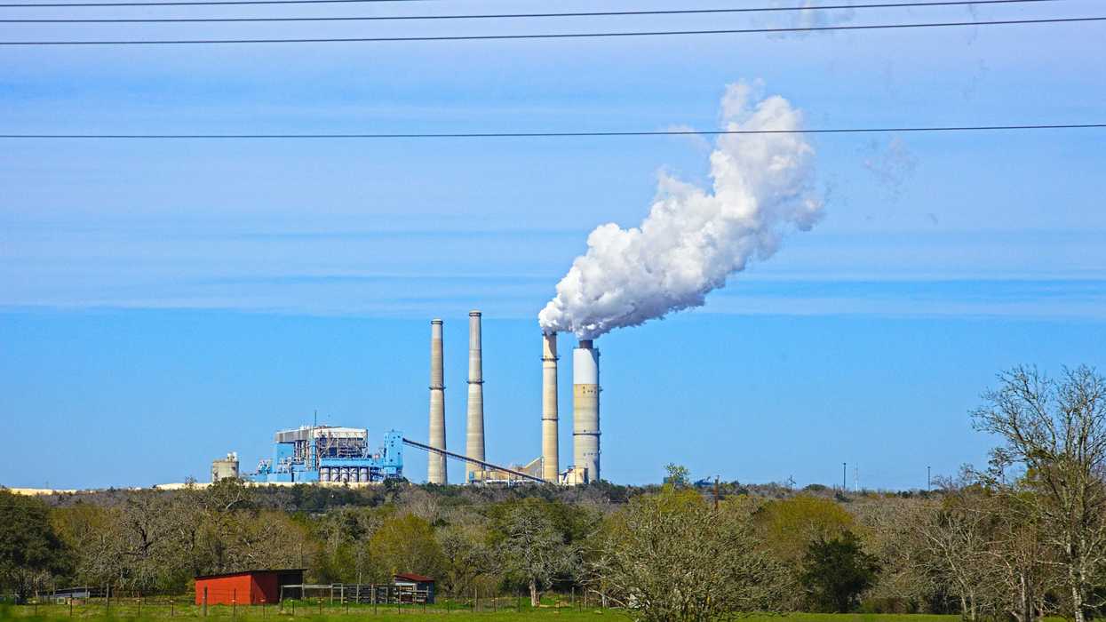 Coal fired power plant with smoke stacks spewing on a hill and a green field in foreground.