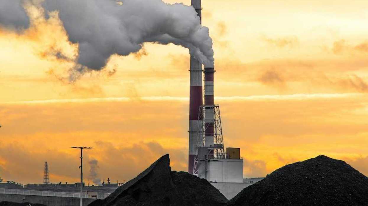 Coal plant emitting smoke from red and white smokestacks adjacent to large piles of coal and coal ash.