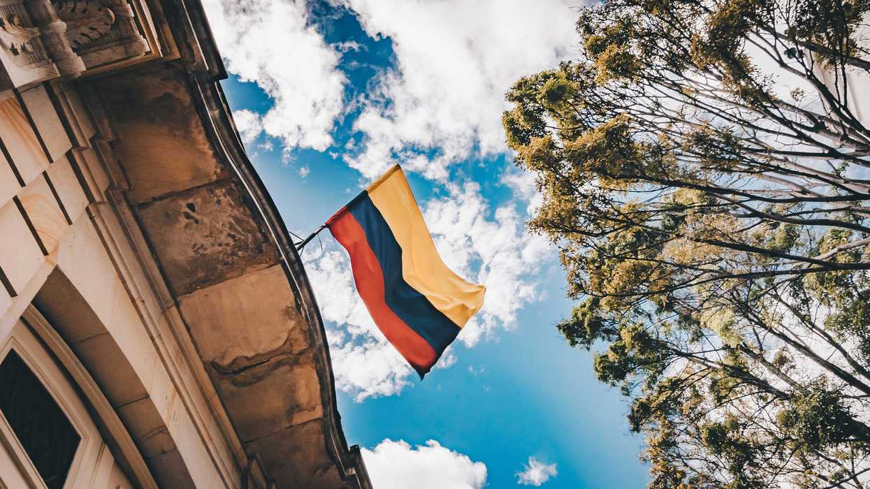 Colombian flag flying from the top of a building as seen from below with clouds and trees in background.
