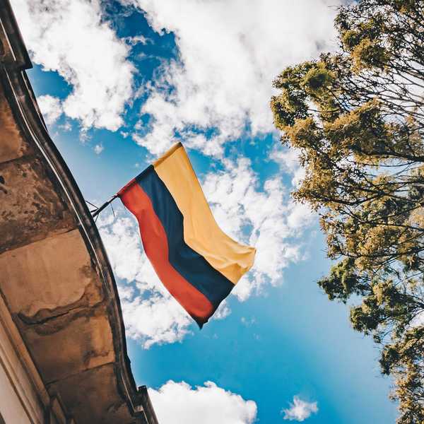 Colombian flag flying from the top of a building as seen from below with clouds and trees in background.