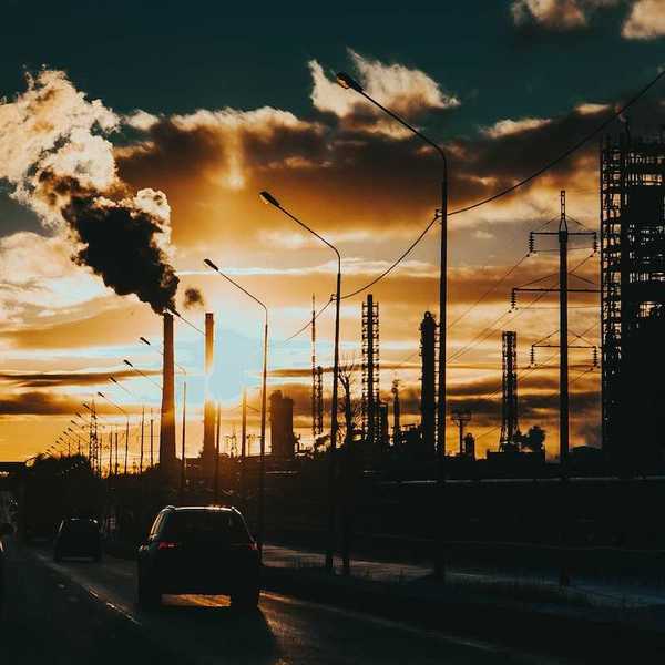 Commuters on an urban expressway beneath belching smokestacks.