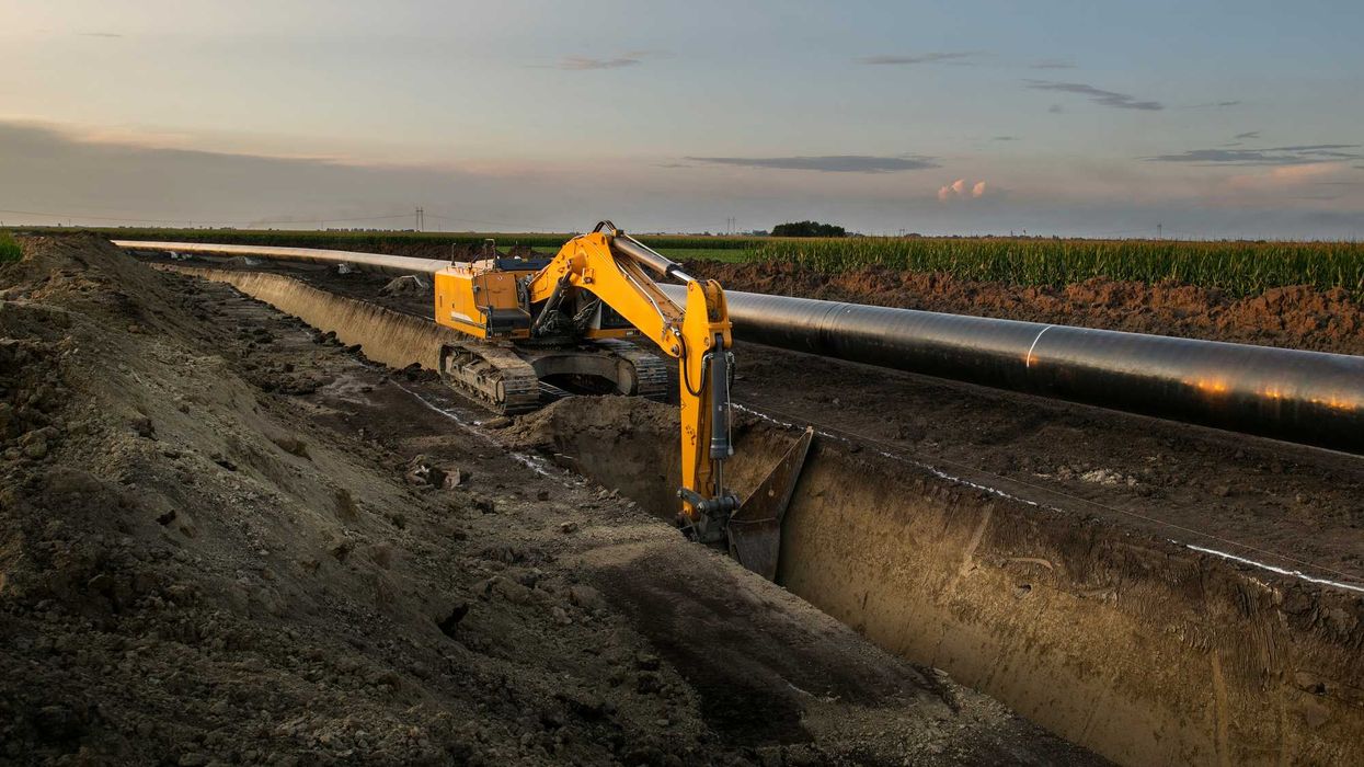 Construction equipment digs a trench along an oil pipeline in a field.