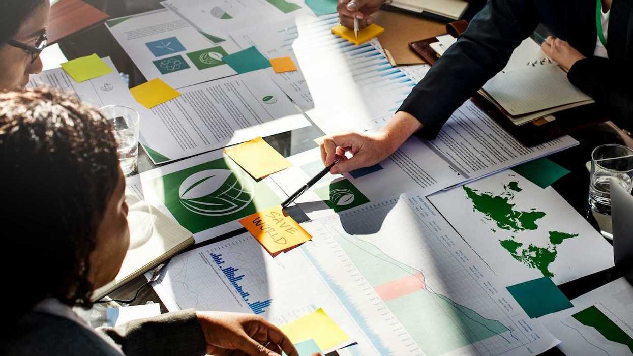 Corporate types gathered around desk full of paperwork with post-it note in center reading "SAVE WORLD."