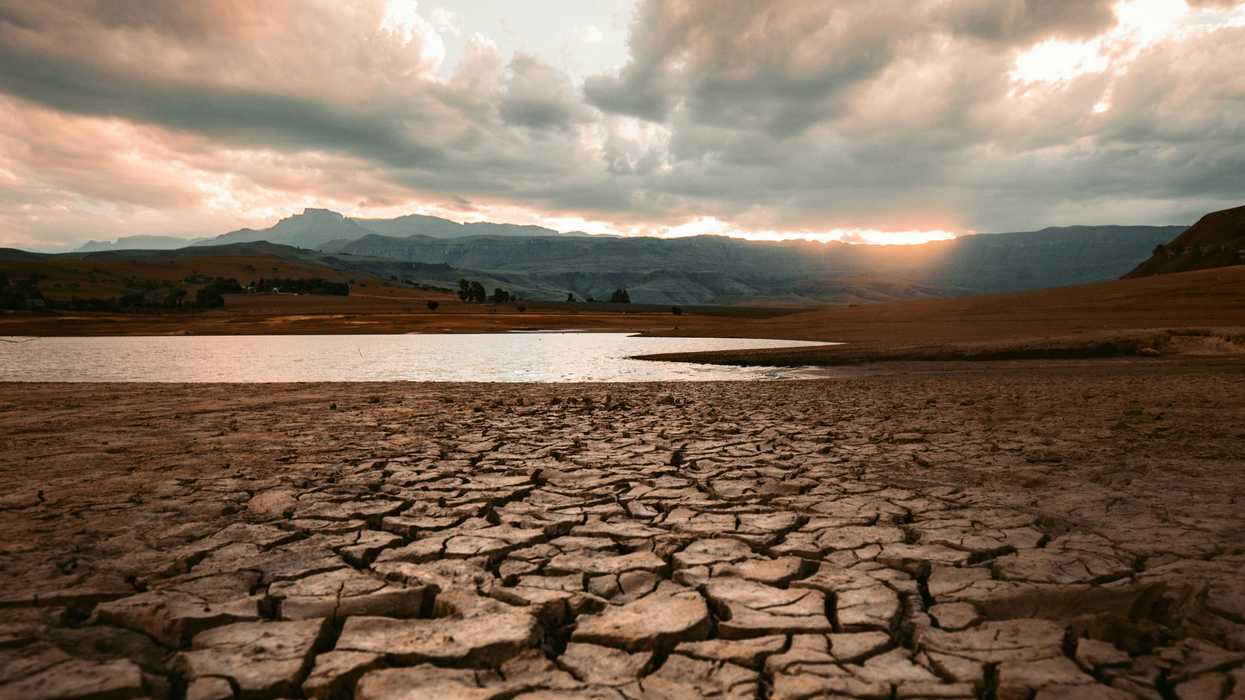 cracked brown dry soil on edge of drought-stricken water body with setting sun and clouds in background.