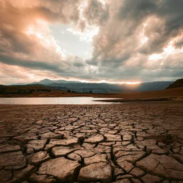 Cracked drought-ravaged foreground, receding water level of lake in background, sun rising or setting on distant hills