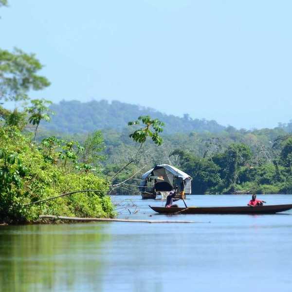 Dugout canoe with two passengers on a river in a rainforest/jungle setting.