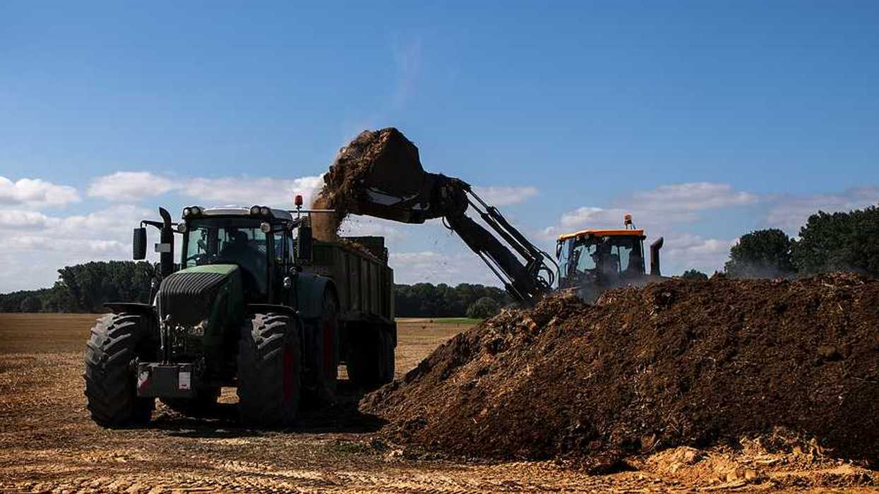 Excavator with a bucket loads organic fertilizer or manure on a tractor-pulled spreader in preparation for application to fields. blue sky copy space