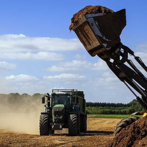 Excavator with a shovel full of manure ready to fill a manure spreader against a partly blue sky