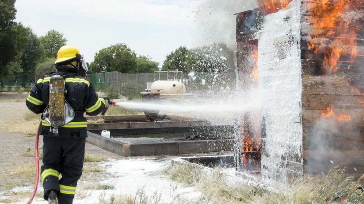 Firefighter dressed in black and yellow spraying firefighting foam (AFFF) on burning structure