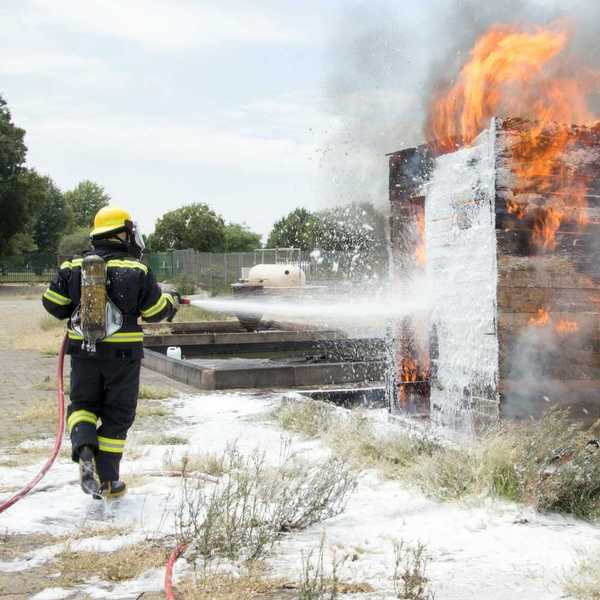 Firefighter dressed in black and yellow spraying firefighting foam (AFFF) on burning structure