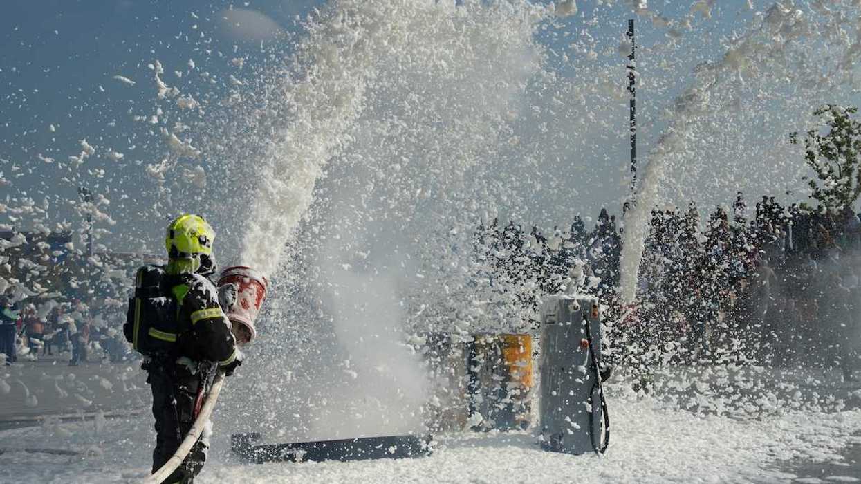 Firefighter in turnout gear covering the surrounding area in toxic firefighting foam during training exercise.